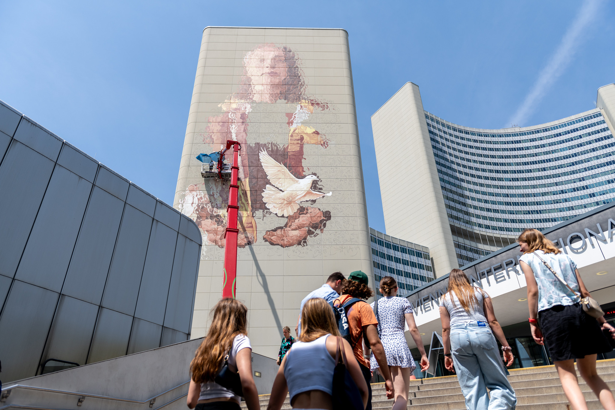 People walking up the stairs with a tall tower behind them and partially painted wall showing woman holding dove.