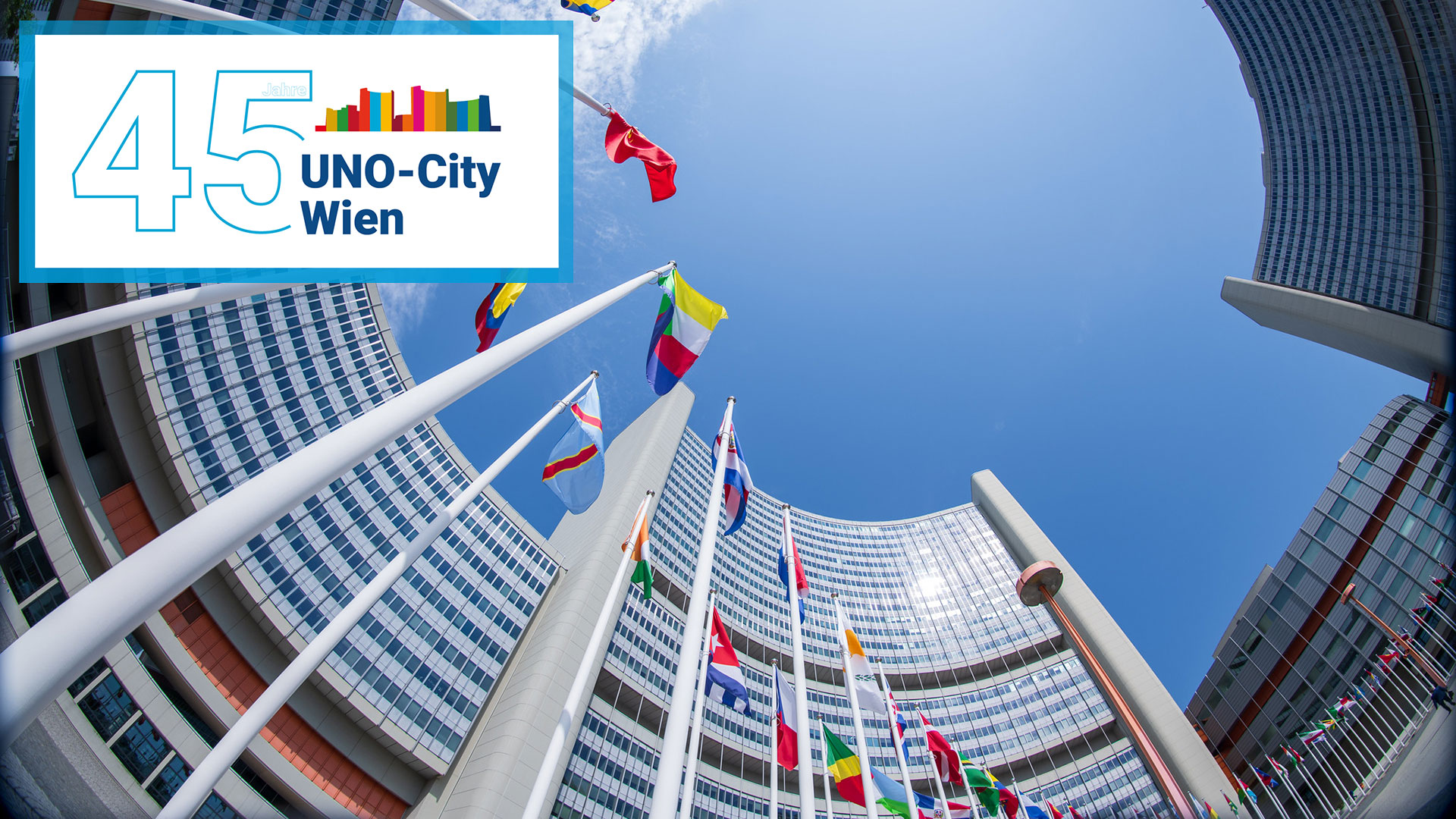 Frog-eye view of the tops of three towers of a building with some flags flying in front of them, with the logo of the 45th anniversary of the Vienna International Centre in the corner.